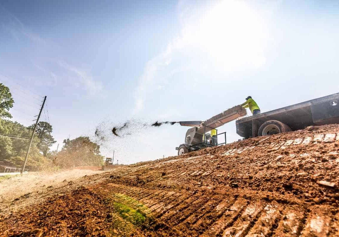 Straw blower spraying mulch across a freshly graded red clay embankment